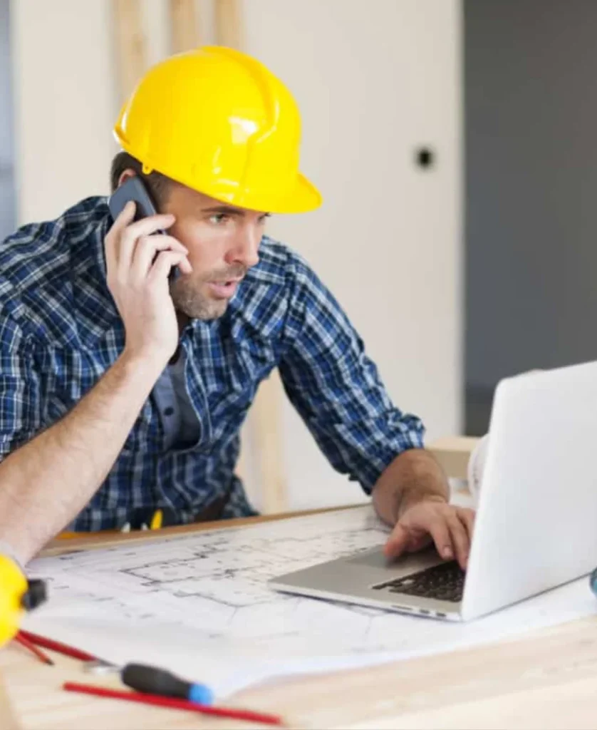 Builder in a hard hat on the phone while checking plans on a laptop